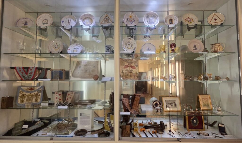 Glass-front display cabinet in a museum, filled with decorative plates on the top shelves, porcelain cups, textiles, framed photos, and small vintage artifacts arranged across multiple glass shelves.