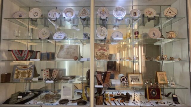 Glass-front display cabinet in a museum, filled with decorative plates on the top shelves, porcelain cups, textiles, framed photos, and small vintage artifacts arranged across multiple glass shelves.