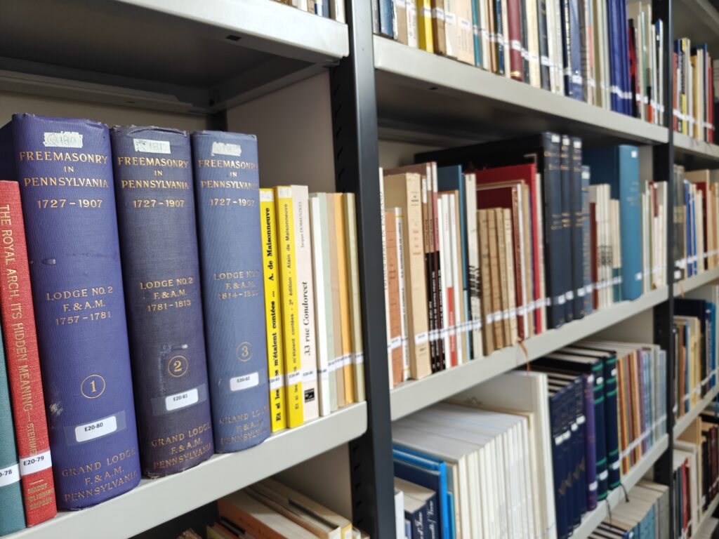 Blue hardcover volumes titled 'Freemasonry in Pennsylvania 1727–1907' on a library shelf, with other colored books nearby on metal shelving.