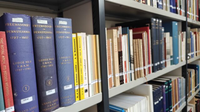 Blue hardcover volumes titled 'Freemasonry in Pennsylvania 1727–1907' on a library shelf, with other colored books nearby on metal shelving.